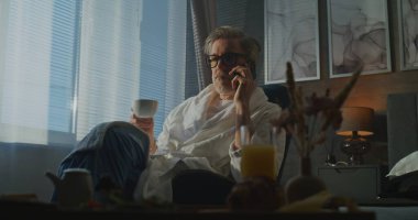 Otel odasındaki bornozda uzaktan kumandayla çalışan Yönetici Sabahı. Kol sandalyesinde oturan, telefonla konuşan, odada dinlenen gri saçlı adam. Bütünleşik İş ve Kişisel Yaşam kavramı.