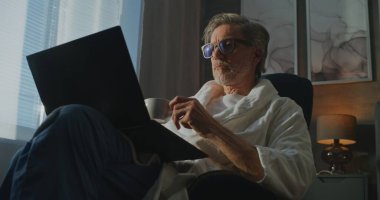 Beyaz Bornozlu Şık Gri Saçlı Adam dizüstü bilgisayardaki e-postaları kontrol ediyor, Otelin Sabah Aydınlığı Atmosferinin tadını çıkarıyor, ve Aroma Coffee. Konfor, İş, Yavaş İş Seyahati Arasındaki Denge.