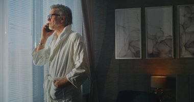 Beyaz Otel Bornozlu Adam Sunny Window 'un yanında duruyor. Otel odasında telefon bekliyor. Uzaktan Çalışma Yaşam Stili, İş ve Eğlence Seyahatinin Birleştirilmesi ve Yolda Profesyonel Bağlı Kalmak.