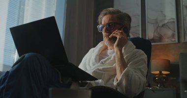 Smartphone 'da Bornozlu Profesyonel Adam konuşuyor ve Sunlight' ta dizüstü bilgisayar kullanıyor. Modern Otel Süitinde Çalışma ve Rahatlama Dengesi. Seyahat ederken Dijital Bağlantı kavramı. El bilgisayarı.