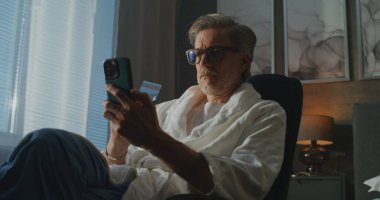 Cüppeli Gri Saçlı Adam Pencere kenarında oturuyor, İnternet üzerinden Telefon ve Kredi Kartıyla Alım Onaylıyor. Modern Teknoloji, Uzaktan Çalışma, Dijital Finans ve Rahat Otel Hayatı kavramı. El bilgisayarı.