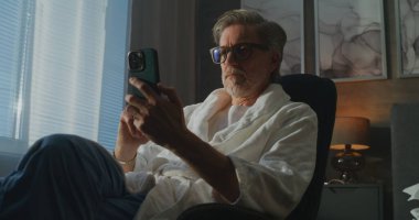 Cüppeli Gri Saçlı Adam Pencere kenarında oturuyor, İnternet üzerinden Telefon ve Kredi Kartıyla Alım Onaylıyor. Modern Teknoloji, Uzaktan Çalışma, Dijital Finans ve Rahat Otel Hayatı kavramı. El bilgisayarı.