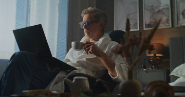 Bornozlu Adam, Pencere kenarında Laptop ve Kahvaltıda Oturuyor, Huzurlu Sabah Parıltısı Üzerine Odaklandı. Üretken Sabahlar, Uzaktan Çalışma ve Zarif Dijital Yaşam Tarzı kavramı. El bilgisayarı.