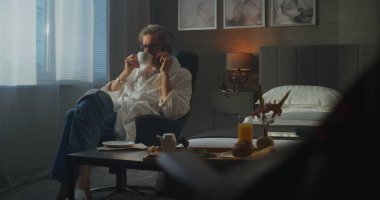 İşadamı Sabah Rutin Telefon Görüşmesi ve Otel Odasında Kahvaltı. Koltuğunda oturan adam telefonda konuşuyor ve kahvaltı tepsisi hazırda bekliyor. Sabah İş Rutini kavramı. Doli Vuruşu.
