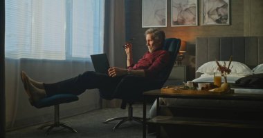 Gri Saçlı Adam Otel Sandalyesinde Rahatça Oturur, Sabah Işığında Dizüstü Bilgisayarda Yazar. Olgun Adam kasıtlı olarak çalışır, Kahvaltıyı unutur. Uzaktan Çalışma, İş Gezileri ve Dijital Esneklik Konsepti.