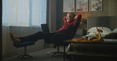 Gri Saçlı Adam Otel Sandalyesinde Rahatça Oturur, Sabah Işığında Dizüstü Bilgisayarda Yazar. Olgun Adam kasıtlı olarak çalışır, Kahvaltıyı unutur. Uzaktan Çalışma, İş Gezileri ve Dijital Esneklik Konsepti.