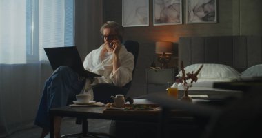 Bornozlu zarif gri saçlı adam otel odasından iş görüşmesini yönetiyor, laptop ve kahvaltı tepsisiyle birden fazla iş yapıyor. Melez Çalışması, Üretkenlik, Seyahat Hayatı Konforu. Doli Vuruşu.