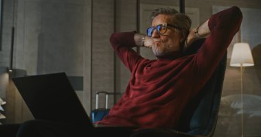 Gri Saçlı Adam Yatakta Oturuyor, Otel Odasının Akşam Işığı 'nda dizüstü bilgisayarla çalışıyor. Kucağında kişisel bilgisayarla koltukta oturan adam. Seyahat, Uzaktan Çalışma, Modern Profesyonel Bağlılık Konsepti.