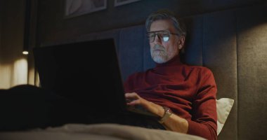 Kişisel bilgisayarı olan son sınıf öğrencisi yumuşak ışıklı modern otel odasında yatağında rahatça oturuyor. Seyahat Kavramı, Çevrimiçi İletişim, Dijital Bağlantı, Modern Uzaktan Çalışma ve Küresel İş Hayatı.