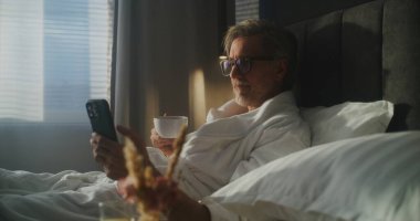 Otel Yatağında Olgun Yönetici Oturur, Bornoz Giyer, Kahvaltı İçin Kahve fincanı tutarken Smartphone Bakar. İş Hayatı Bütünleşmesi, Mobil Teknoloji Kullanımı, İş Gezisi. Doli Vuruşu.