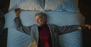 Cozy Otel Odası 'ndaki Beyaz Yatak' a Mutlu Yıllar Giyen Adam düşüyor. İş seyahatinden yorgun düşen adam yatağında dinleniyor. Otelde Yaşam Kavramı, Gezgin Yorgunluğu, İş Gezisi. Üst Görünüm.