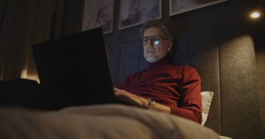 Dark Hotel Room 'da dizüstü bilgisayara odaklanmış adam çalışıyor. Olgun Adam Yatağında Rahat Oturuyor, Yatağın başındaki lambalarla aydınlatılıyor. Sorumluluk sahibi çalışma, uzaktan çalışma ve gece yarısı verimliliği kavramı. El bilgisayarı.