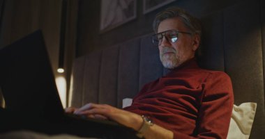 Stylish Otel Odasında Dizüstü bilgisayarda Yaşlanmış İşadamı Çalışıyor. Loş Işıklandırma ve Sıcak Atmosfer 'de, Yataktaki Adam Kişisel Bilgisayarı Kullanıyor. Modern Uzaktan Çalışma ve Küresel İş Hayatı kavramı. El bilgisayarı.