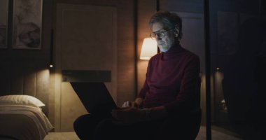 Dimly Lit Otel Odası 'nda dizüstü bilgisayarda gözlük takan odaklanmış bir adam. Gri Saçlı Adam Kişisel Bilgisayarı kullanarak Gece Çalışıyor. Modern Yaşam Konsepti, Teknoloji Tarafından Sürülen, İş Rutini. Doli Vuruşu.