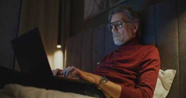 Profesyonel Olgun Adam, Karanlık Otel Odasında Yatakta Otururken Bilgisayarda ve E- postaları Kontrol Ediyor. Konsept Seyahat, Çevrimiçi İletişim, Dijital Bağlantı, Modern Uzaktan Çalışma, Küresel İş Hayatı.