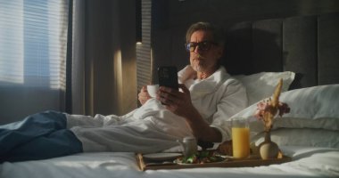 Otel Yatağında Olgun Yönetici Oturur, Bornoz Giyer, Kahvaltı İçin Kahve fincanı tutarken Smartphone Bakar. İş Hayatı Bütünleşmesi, Mobil Teknoloji Kullanımı, İş Gezisi. Doli Vuruşu.