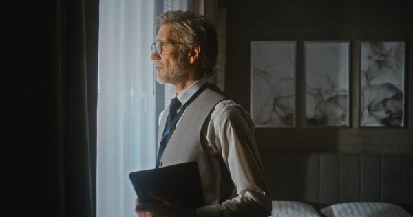 Executive Ready для презентации в номере отеля "Солнечный". Уверенный в себе бизнесмен стоит под солнцем у окна, держа в руках цифровой планшет, готовый к встрече. Обучение, цифровой рабочий процесс. Портрет.