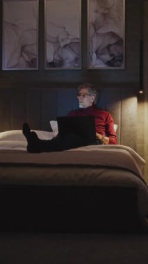 Balıkçı yaka yakalı yakışıklı adam Dimly Lit 'te, Modern Otel Odasında, yatağında otururken dizüstü bilgisayarda çalışıyor. Sessiz konsantrasyon ve yalnızlık. Uzaktan Kumanda, İş Gezisi, Dijital Çağ. Dikey Çekim.