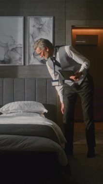 Resmi Takım Elbise Denetleme Odası 'nda Zarif Olgun Otel Yöneticisi Tablet Fotoğrafı Kullanıyor ve Yatak Düzenlemelerini Değerlendiriyor. Konukseverlik Standartları, Kalite Kontrol, Profesyonel Yönetim. Dikey Çekim.