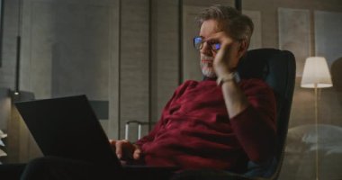 Gri Saçlı Adam Yatakta Oturuyor, Otel Odasının Akşam Işığı 'nda dizüstü bilgisayarla çalışıyor. Kucağında kişisel bilgisayarla koltukta oturan adam. Seyahat, Uzaktan Çalışma, Modern Profesyonel Bağlılık Konsepti.