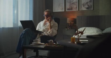 Bornozlu zarif gri saçlı adam otel odasından iş görüşmesini yönetiyor, laptop ve kahvaltı tepsisiyle birden fazla iş yapıyor. Melez Çalışması, Üretkenlik, Seyahat Hayatı Konforu. Doli Vuruşu.