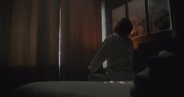 Pijama ve bornoz giyen Olgun Adam Yataktan Kalkar ve Kalın Perdeleri Arkaya Çeker, Güneş Işığını Odaya Aldırır. Adam kaslarını esnetir ve gerer. Rahatlamayı, seyahati, duygusal refahı kabul et.