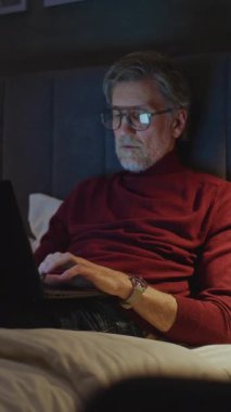 Gözlüklü, zarif, olgun bir adam yatakta otururken dizüstü bilgisayar kullanıyor. Sıcak Akşam Işığı ve Sakin Atmosferinde, İnsan kişisel bilgisayarı üzerinde çalışır. Konsept Seyahat, Dijital Bağlantı. Dikey Çekim.