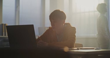 Teenage Student, Sunlit Sınıfı 'ndaki laptopta sessizce çalışıyor. Sınıf arkadaşı telefondaki mesajları kontrol ediyor. Modern Çalışma Ortamı, Dijital Öğrenme Alışkanlıkları kavramını. Yavaş Hareket.