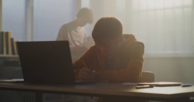 Teenage Boy, Soft Afternoon Light 'ta Masa Oturuyor, Laptop ekranına odaklanmış. Arka planda, telefonda başka bir öğrenci parşömeni var. Eğitimde Dijital Teknoloji kavramı, Dijital Alışkanlıkları Vurguluyor.