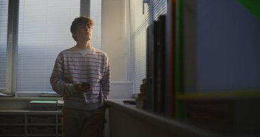 Çizgili Süveterli Genç Ders Salonunun Parlak Penceresinde Bekliyor, Telefonu Kontrol Ediyor, Dikkatlice Dışarıya Bakıyor. Öğrenci Yansıması, Dijital İletişim, Modern Kampüs Hayatı.