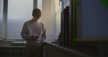 Çizgili Süveterli Genç Ders Salonunun Parlak Penceresinde Bekliyor, Telefonu Kontrol Ediyor, Dikkatlice Dışarıya Bakıyor. Öğrenci Yansıması, Dijital İletişim, Modern Kampüs Hayatı.