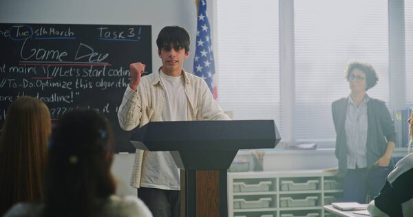 Молодой студент стоит на пьедестале почета и произносит страстную речь перед одноклассниками в современном аудитории на фоне американского флага, используя мощные, энергичные жесты. Лидерские качества студентов.