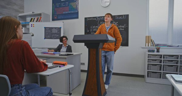 Студент в оранжевой толстовке стоит за подиумом перед одноклассниками и уверенно говорит, используя выразительные выражения лица и жесты. Концепция ораторских навыков и интерактивного обучения.
