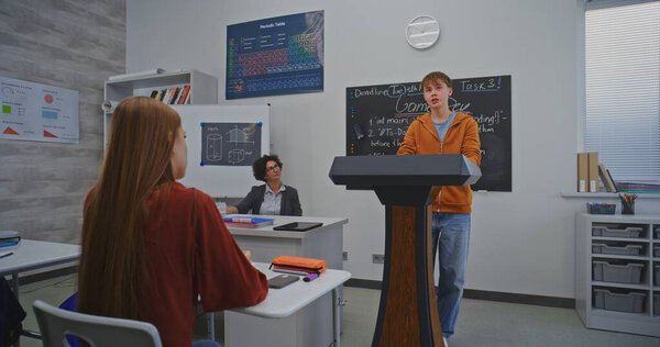 Окаменелый мальчик в ярко-оранжевом худи проводит классную презентацию проекта "Творческая школа". Учитель и ученики слушают внимательно. Творчество, творчество и коллективизация в образовании.