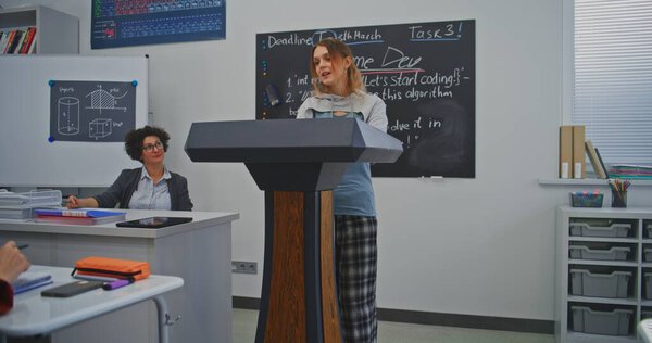 Молодая женщина уверенно говорит на подиуме, улыбаясь, обращаясь к одноклассникам и учителю. Концепция лидерства, уверенности и роста коммуникационных навыков через современное образование. Долли Шот.