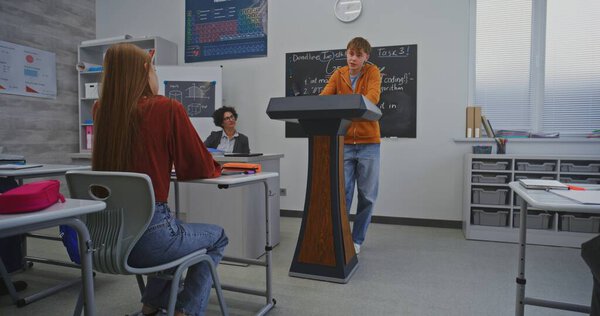 Студент в оранжевой толстовке стоит за подиумом перед одноклассниками и уверенно говорит, используя выразительные выражения лица и жесты. Концепция ораторских навыков и интерактивного обучения.