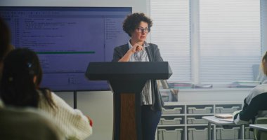 Kadın Kodlama Öğretmeni Podyumda Büyük Sayısal Ekranda Söz Dizimini Açıklıyor. Modern Bilgisayar Bilimi Eğitimi, BTMM Öğrenme, Teknoloji Edebiyatı ve Öğretim Yeniliği kavramını benimse.
