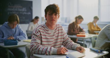 Süveter giyen çocuk masasında sakin ve kendinden emin bir şekilde oturuyor. Sınıf arkadaşları sessizce arkada çalışır. Konsept Sınıfı Atmosferi, Öğrenci İfadesi Destekleyici Öğrenim Ortamı. Ağır çekim. Portre.