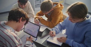 Dört Genç Öğrenci Masa etrafında toplandı, Yoğun Çalışma Oturumuna Odaklandı. Not Defterlerine Aktif Yazan ve Grafik ve Grafikleri Gösteren Dijital Tablet Danışmanlığı Yapan Çocuklar. Sabit Çekim.