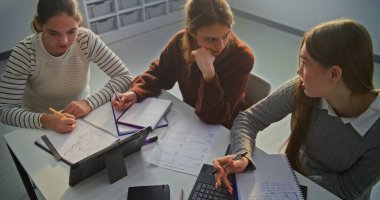 Masada oturan genç kızlar ödev yapıyor, diyagram çiziyor ve birlikte testler yapıyorlar. Modern Sınıfta İşbirliği, Akademik Odaklanma ve Üretken Çalışma Atmosferi Konsepti. Üst Görünüm.