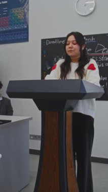 Genç Bayan Öğrenci Çekici Gülümseme ile Podyumda Duruyor, Parlak Sınıfta oturan sınıf arkadaşına rapor veriyor. Masadan Öğretmen Gözlem. Toplum içinde konuşma, iletişim becerileri. Dikey Çekim.