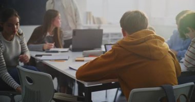 Paylaşılan Çalışma Alanı 'nın etrafında toplanan bir grup öğrenci laptopları, tabletleri ve kâğıt notları kullanarak atamalarda aktif olarak işbirliği yapıyor. Kooperatif problem çözme kavramı ve Modern Sınıf Takım Çalışması.