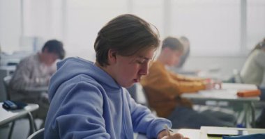 Açık Mavi Kapüşonlu Genç Erkek Öğrenci, Parlak Sınıfta Kağıt Üzerine Yazarken Yoğun Konsantrasyon Ediniyor. Modern Okul Ayarlarında Öğrenci Odaklanması, Bağımsız Çalışmanın Süreci.