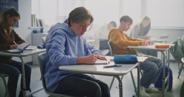 Genç çocuk sınıfta yazma işine odaklanmış durumda. Masada çalışan diğer öğrenciler. Öğrenci Odak Noktası, Akademik Konsantrasyon, Modern Okul Ayarlarında Bağımsız Çalışmalar Süreci. Sabit Çekim.