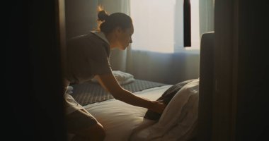 Bir sonraki konuğumuz için üniformalı bir otel görevlisi hazırlanıyor. Kadın perdeleri açar, yatağı düzeltir, bornozu katlar. Konsept Temizlik, Kalite Hizmeti ve Konforlu Seyahat Kalışı.