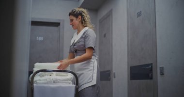 Temiz Üniformalı Kadın Otel Temizlikçisi, Temizlik Trolleyle Yanyana Duruyor, Taze Beyaz Havluları Güzelce Katlıyor. Konsept Temizlik, Düzen ve Yüksek Konukseverlik Hizmeti Standartları.