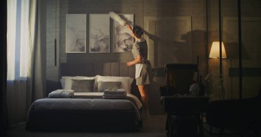 Profesyonel Üniformalı Hizmetçi Otel Odası Duvarındaki Resim Çerçevelerini Nazikçe Temizliyor, Beyaz Tüy Duster ile Tozu Kaldırıyor. Konsept Temizlik, Konukseverlik, Oda Bakımı, Detaylara Dikkat.