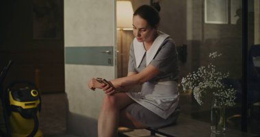 Otel Temizlikçisi Oda Sandalyesinde Kısa Mola Verdi. Temizlik işlerini bitirdikten sonra, Temiz Üniformalı Genç Hizmetçi Hafif Kol Sandalyesine Oturur, Akıllı Telefondaki Mesajları Kontrol eder. İş Hayatı Dengesini Kabul Et.