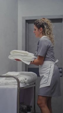 Otel koridorunda kıvırcık saçlı oda hizmetlisi, içerideki servis arabasından temiz havluları dikkatlice katlıyor ve düzenliyor. Modern Konukseverlik Konuk Deneyimi. Dikey Çekim.