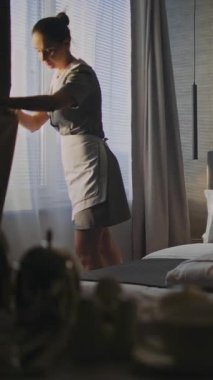 Otel odası personeli perdeleri açar, oda atmosferini tazeler ve mükemmel bir yatak hazırlar. Konsept Konukseverlik, Temizlik, Profesyonel ve Konforlu Seyahat Kalıcılığı. Dikey Çekim.